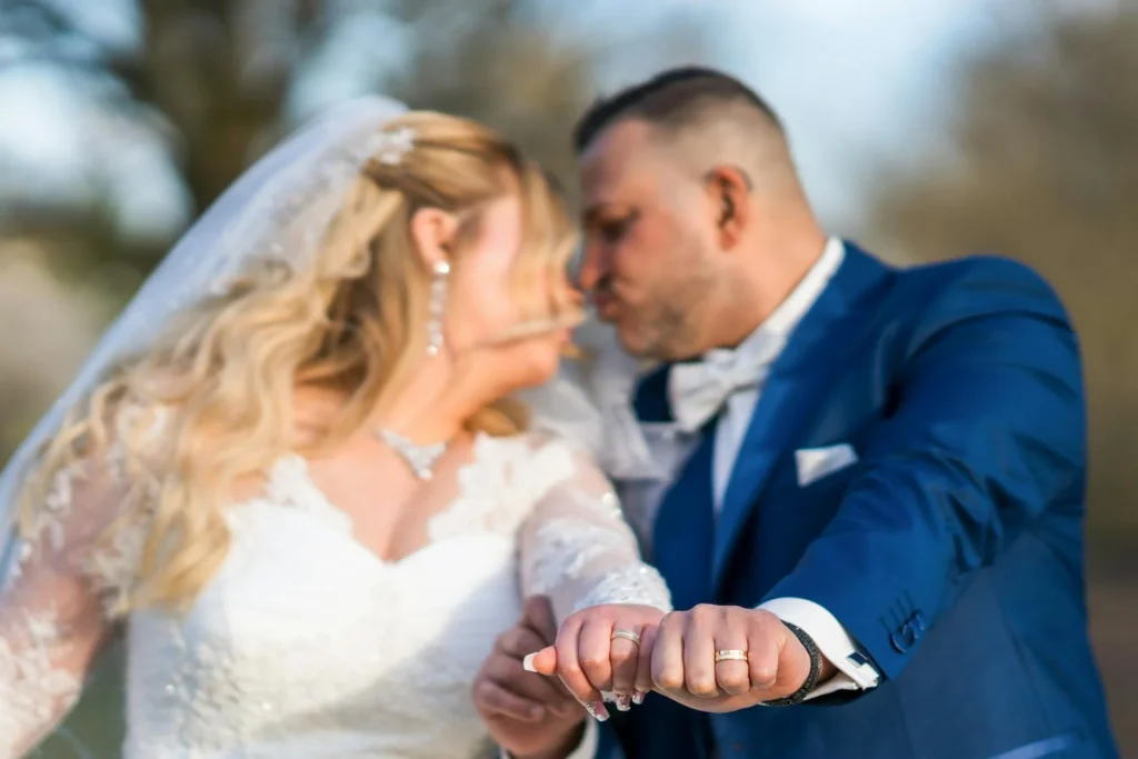 A bride and groom share a kiss while displaying their wedding rings outdoors.