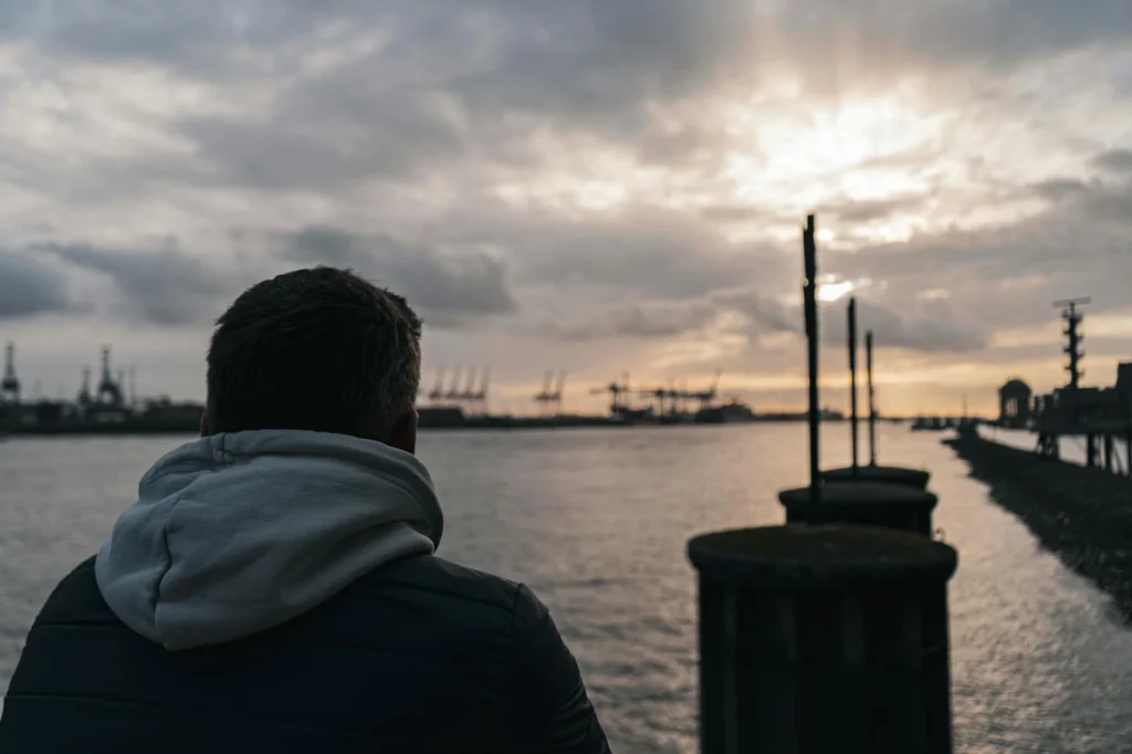 A man reflects by the water with Hamburg's port cranes silhouetted against a sunset.