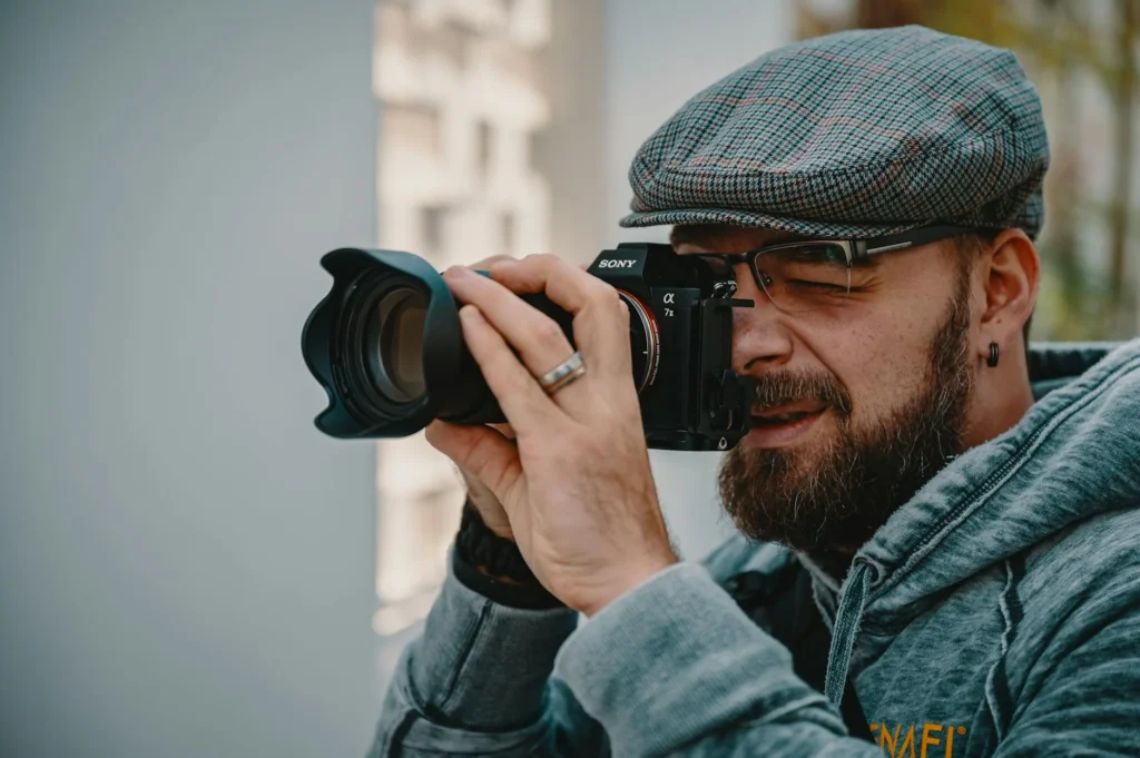 A bearded male photographer using a camera outdoors wearing a plaid cap.